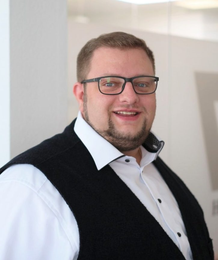 Mann mit Brille, hellem Hemd und dunkler Weste, lächelt freundlich in die Kamera.