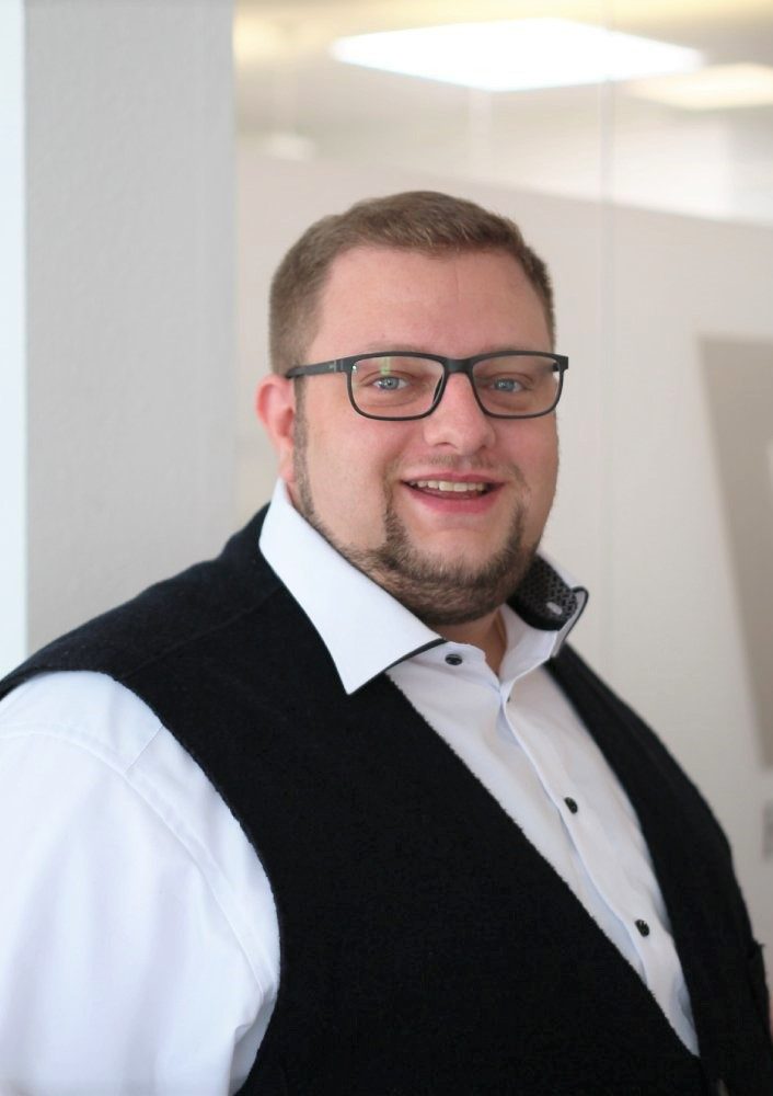 Mann mit Brille, hellem Hemd und dunkler Weste, lächelt freundlich in die Kamera.